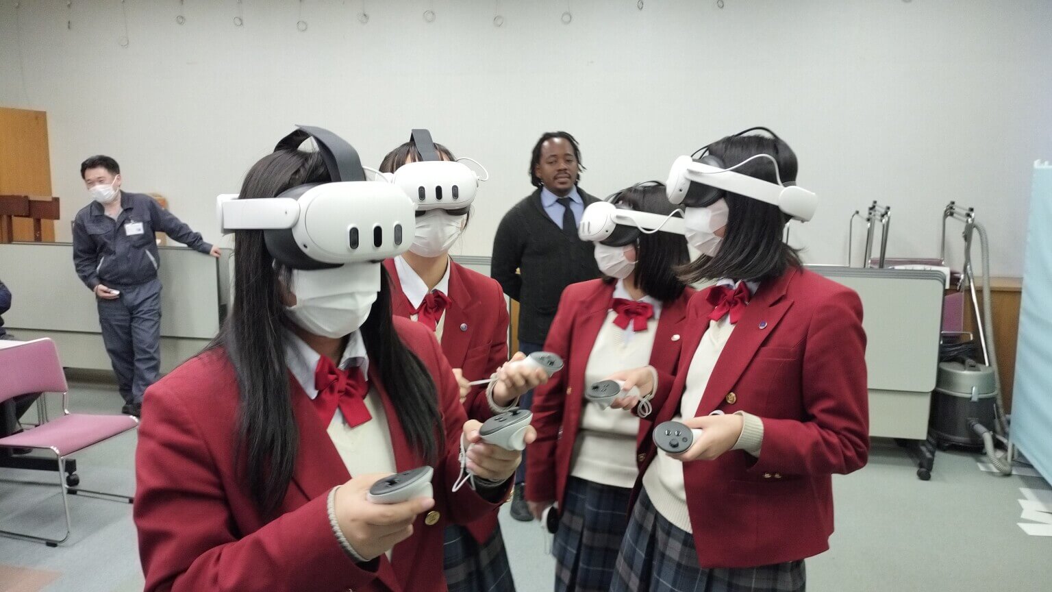 赤いブレザーの女子生徒たちがVRゴーグルとコントローラーでVRChatを体験している様子